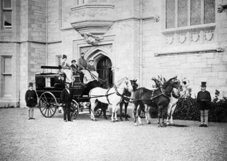 Black horse-drawn carriage with several passengers in front of a light stone building entrance.  Two men stand near the carriage, and a third attends two horses in front of the carriage.