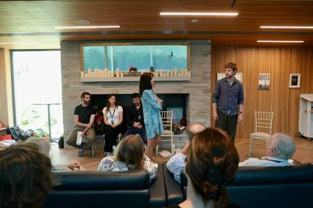 Five people sit and stand before an audience in a room with a wood-paneled wall, stone fireplace, and large window.  A "Roots" poster hangs on the wall.  The audience sits on a dark sofa facing away from the camera.