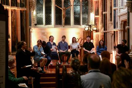 Seven panelists sit on chairs facing an audience in a dimly lit room with marble columns and a crucifix on the wall behind them.  Some panelists hold papers and a woman in a dark top gestures with her right hand.