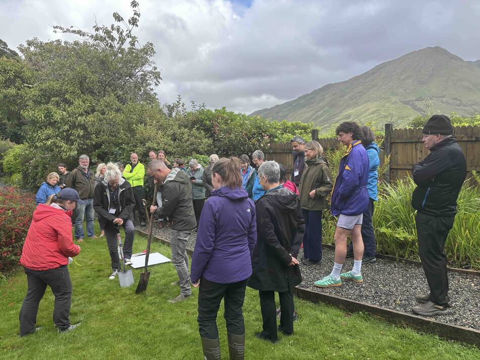 A group of people observe as a woman in a red jacket uses a shovel for a groundbreaking ceremony, with a green mountain in the background.