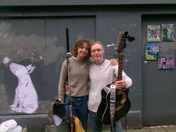 Two people stand against a gray wall with art and posters. A young woman with shoulder-length brown hair, wearing an olive sweater and jeans, smiles at the camera with her arm around a man. He holds an acoustic guitar and wears glasses and a gray long-sleeved shirt.