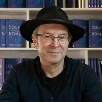 Mike McCormack. An older gentleman wearing a black hat, glasses, and a black shirt, with a multiple copies of the book Solar Bones on a bookshelf behind him.