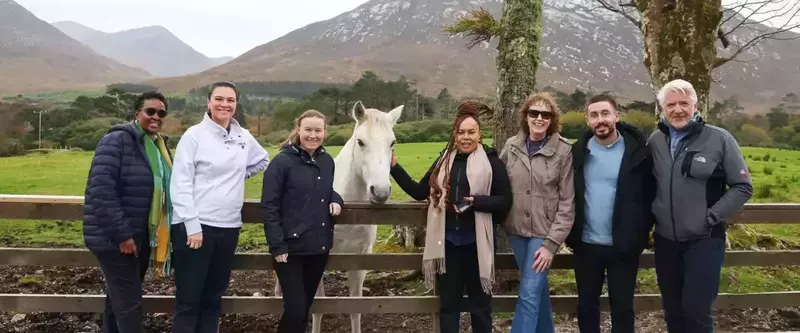 A diverse group of seven people, bundled in jackets and casual outdoor wear, stand together for a photo in front of a light-colored horse. They are posing outdoors on a grassy field, behind a wooden fence. In the background, there are large, rolling, brown and green mountains under a cloudy sky.
