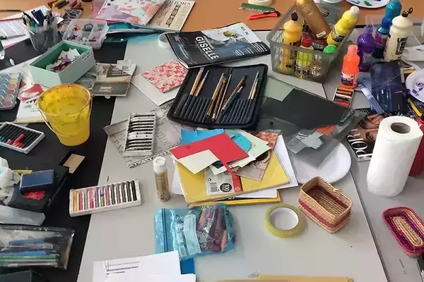 A large wooden table is completely covered with diverse art and craft supplies: colorful paints, brushes, paper, magazines, crayons, glue, and small containers, indicating an active creative workspace.