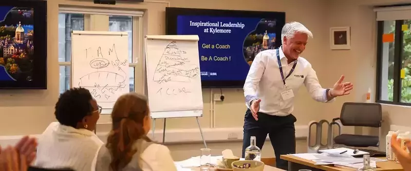 A smiling man in a white shirt gestures while presenting to two workshop attendees. A screen shows Notre Dame's Golden Dome and "Inspirational Leadership at Kylemore" from Notre Dame Global.