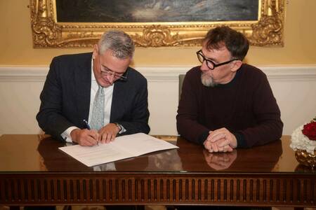 A man in a blue suit signs a document with a blue pen on a polished wooden desk. Another bearded man in a dark sweater and glasses watches intently.