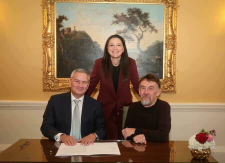 Two men and one woman smile while posing formally at a dark wood table. The man on the left, in a suit, rests his hands on documents. A bearded man sits on the right. A woman in a dark red suit stands centered behind them.