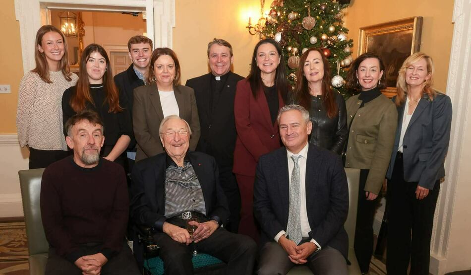 Twelve smiling individuals, nine standing and three seated, pose indoors. A priest in black stands at the back. An older man seated front-center, wearing glasses and a patterned shirt, broadly smiles while holding a champagne glass. A festive Christmas tree is in the background.