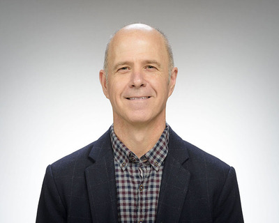 James Lang. A smiling middle-aged bald man wears a dark blue blazer over a red, blue, and white plaid shirt, against a light gray background.