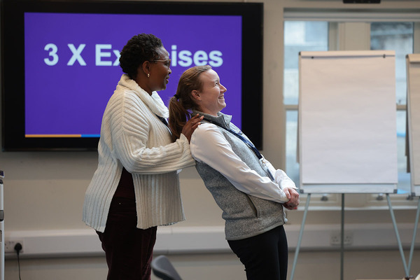 Women in Leadership Program trust exercises. A Black woman in a cream sweater gently supports a smiling white woman in a grey vest who leans back in a stretch. A screen displays "3 X Exercises" in the background.