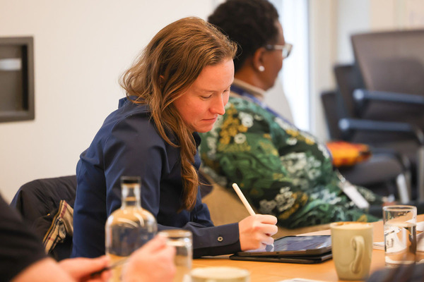 Women in Leadership Program courses. A woman with long brown hair in a navy blue shirt leans forward, focused on writing with a stylus on a tablet at a wooden table. A blurry figure sits in the background.
