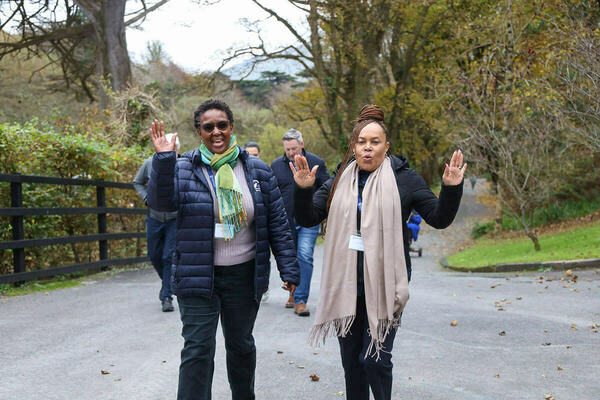 Women in Leadership Program excursions. Two women, smiling brightly and waving, walk on a paved outdoor path. The woman on the left wears a blue puffer jacket and green scarf. The woman on the right wears a black jacket, beige scarf, and has a surprised expression. Autumn trees and a fence are behind them.