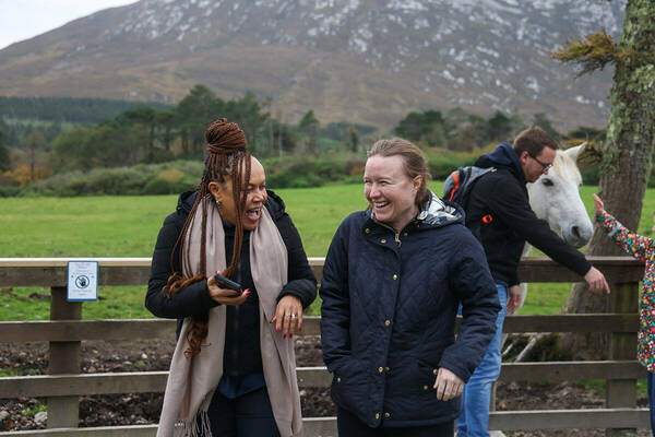 Women in Leadership Program excursions. Two women laugh heartily outdoors. One wears a black coat & tan scarf; the other a navy quilted jacket. A man, white horse, and child interact nearby, with green fields and a mountain in the background.