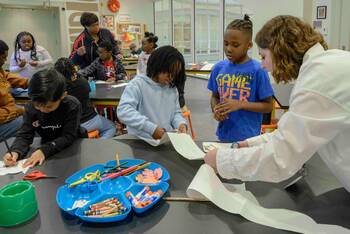 A group of children and adults engage in an art activity at a long dark table. Crayons, glue, and paper cutouts fill a blue craft tray.
