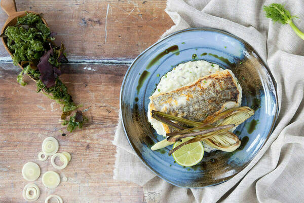 Pan-seared fish with crispy skin rests on creamy white risotto, garnished with roasted fennel and a lime wedge on a blue ceramic plate. A wooden spoon holds green and red seaweed on a rustic wooden table, with sliced raw leeks.