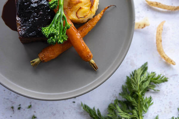 A plate holds a rich, dark braised meat with sauce, a golden potato gratin, two roasted orange carrots, and vibrant green broccolini. Raw potatoes, fresh carrot greens, garlic, and thyme are on the light background.