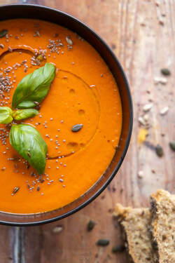 Vibrant orange tomato soup in a dark bowl, garnished with fresh green basil leaves, a drizzle of olive oil, flax, chia, and pumpkin seeds. Slices of multigrain bread rest on a rustic wooden table.