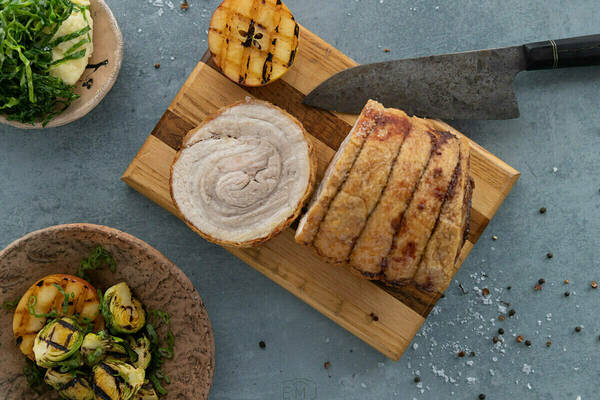 Golden-brown porchetta roast and slice on a wooden board next to a chef's knife. Accompanied by grilled apple, roasted Brussels sprouts, and mashed potatoes with greens.