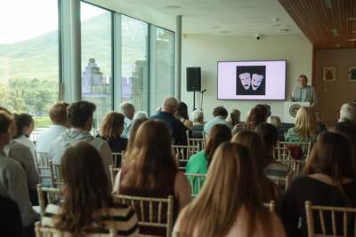 Man speaks at Notre Dame podium, theatrical masks on screen. Kylemore Abbey and mountain visible through window.