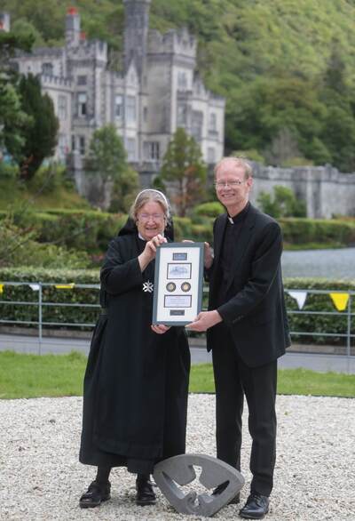 Smiling nun and Rev. Robert K. Dowd, C.S.C., holding framed plaque outside Kylemore Abbey with green hills.