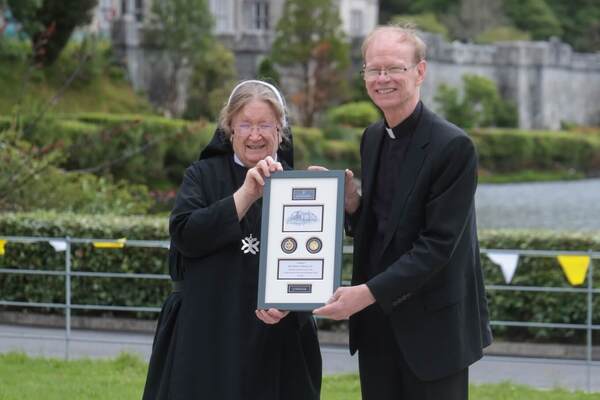 Smiling nun and Rev. Robert K. Dowd, C.S.C., holding framed plaque outside Kylemore Abbey with green hills.