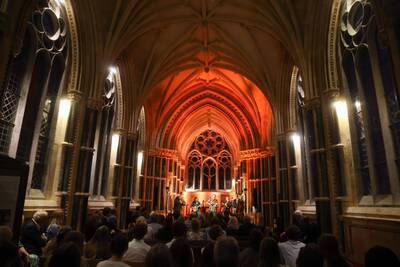 Musicians perform on stage in a grand Gothic church with a rose window and orange-lit arched ceiling for a seated audience.