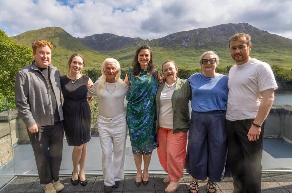 Seven people smile while posing on an outdoor patio, with green mountains, a lake, and a cloudy sky behind them.